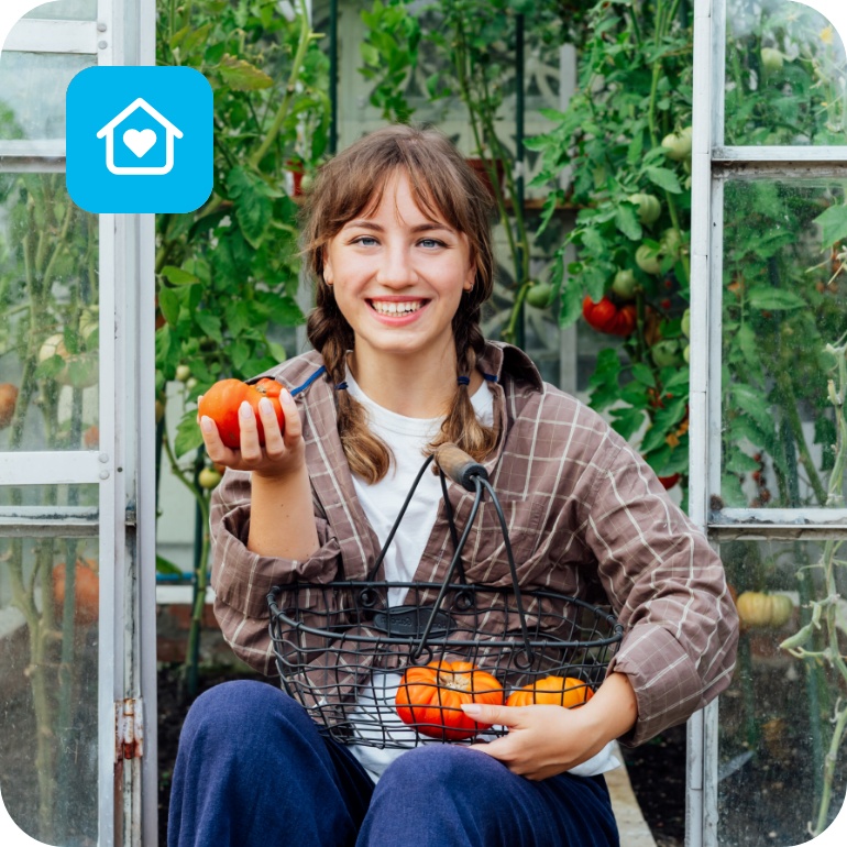 Frau sitzt im Gewächshaus und hält eine frisch geerntete Tomate und einen Korb mit weiteren Tomaten.