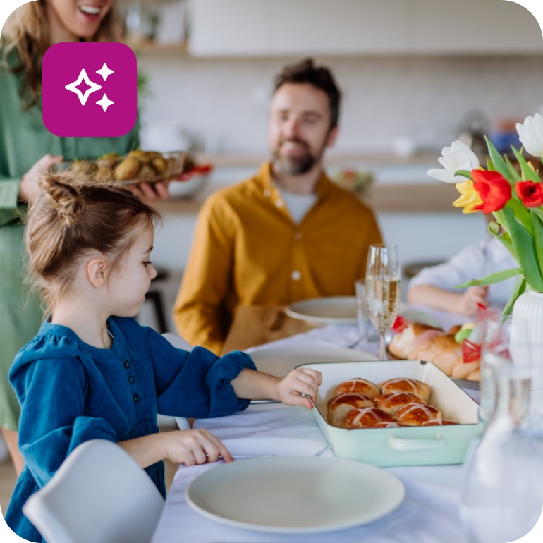 Eine Familie sitzt bei einem Osterbrunch beisammen. Auf dem Tisch stehen Tulpen und Ostergebäck wie süße Brötchen und ein Hefezopf.