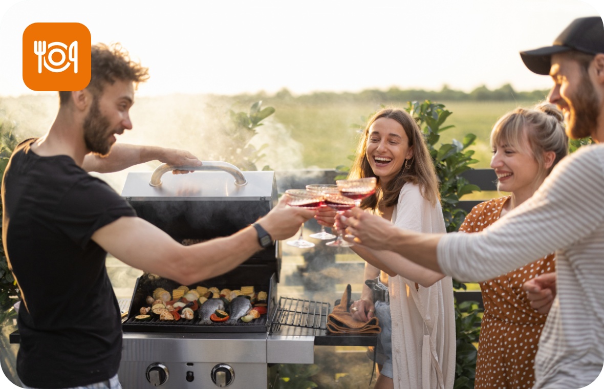 Vier Personen stoßen auf einer Terrasse neben einem Gasgrill stehend mit Rotwein an.