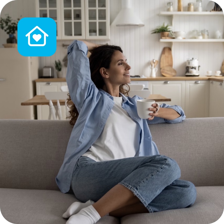 Frau sitzt entspannt auf einem Sofa mit einer Tasse Kaffee in einer hellen aufgeräumten Wohnküche.