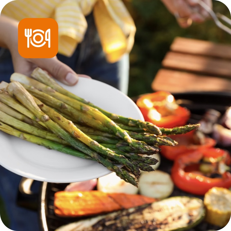 Gegrillter grüner Spargel auf einem Teller. Im Hintergrund verschiedenes gegrilltes Gemüse auf dem Grill.