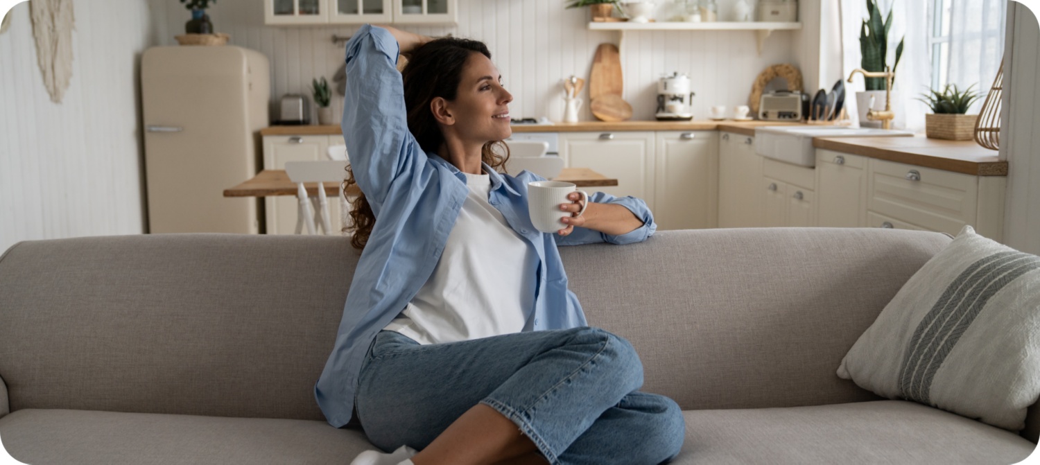 Frau sitzt entspannt auf einem Sofa mit einer Tasse Kaffee in einer hellen aufgeräumten Wohnküche.