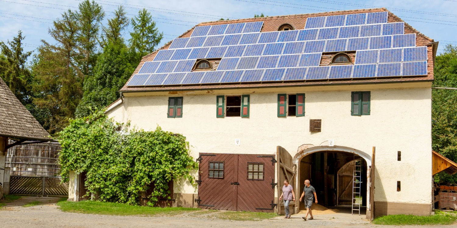 Ein Bauernhofgebäude mit Solarpanels auf dem Dach, umgeben von grüner Natur, zwei Personen gehen durch das Tor.