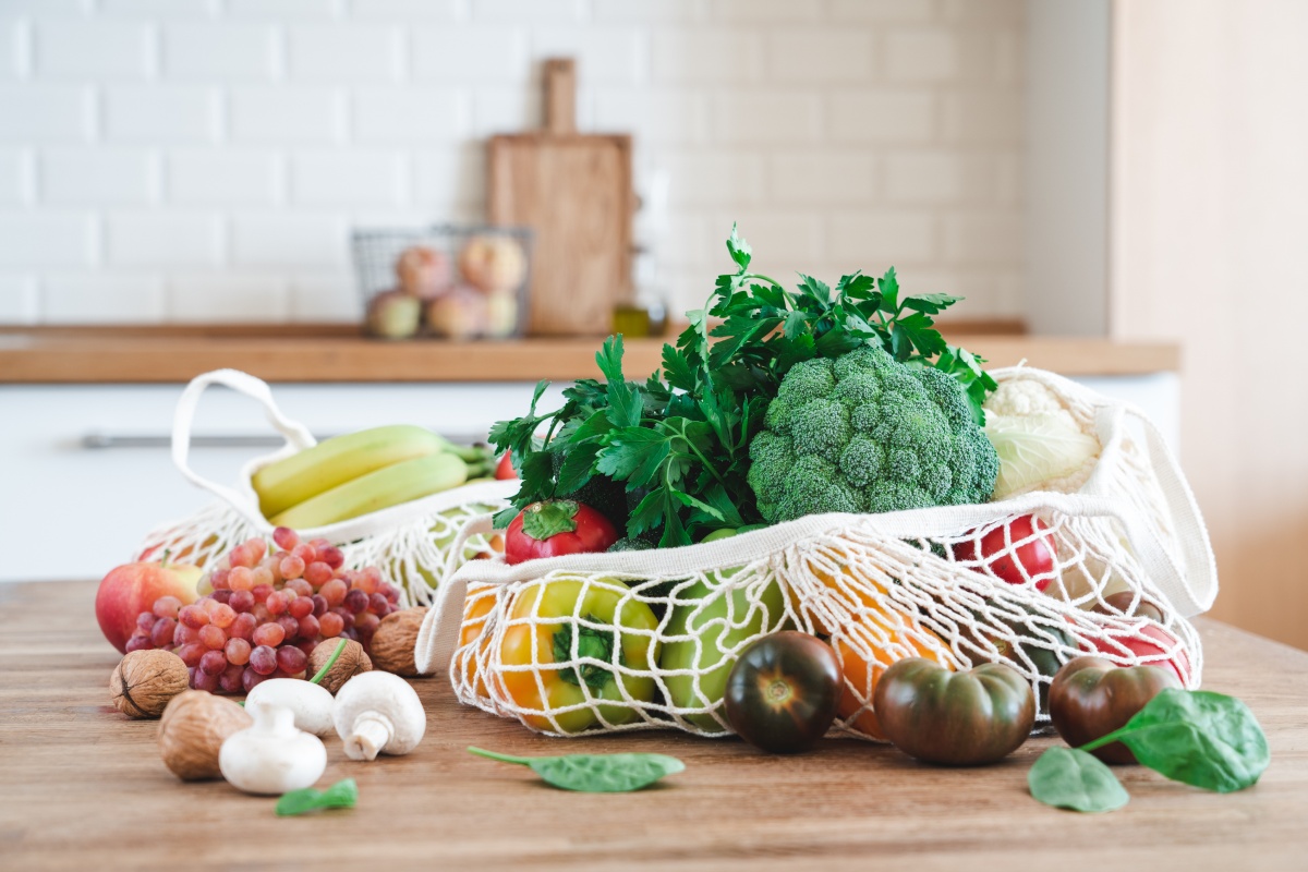 Netztasche gefüllt mit frischem Obst, Gemüse und Kräutern, auf einem Holztisch in einer modernen Küche.
