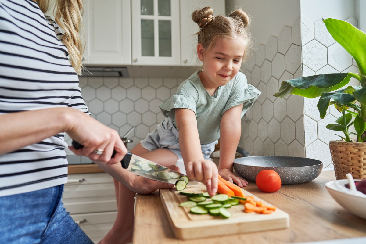 Mutter schneidet Gemüse, während ihre Tochter auf der Küchentheke sitzt und Gurkenscheiben vom Schneidebrett nimmt.