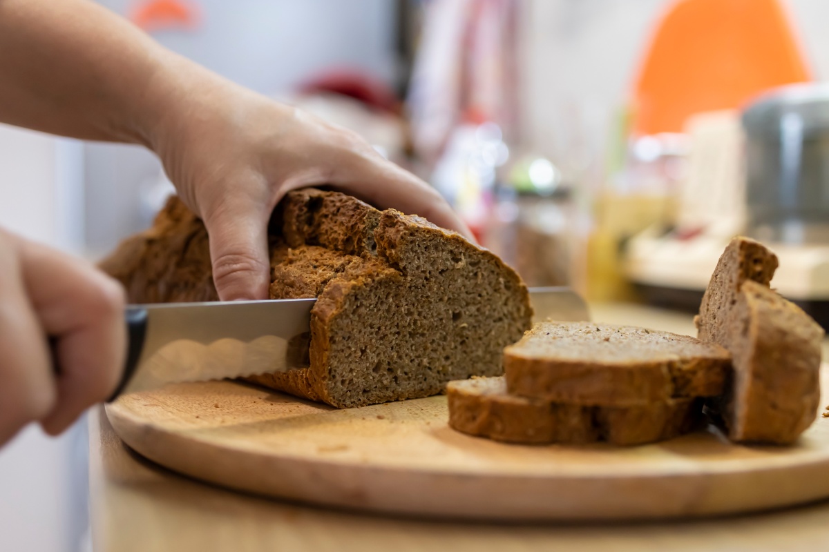 Person schneidet frisches Vollkornbrot auf einem Holzbrett mit einem Brotmesser in dicke Scheiben.
