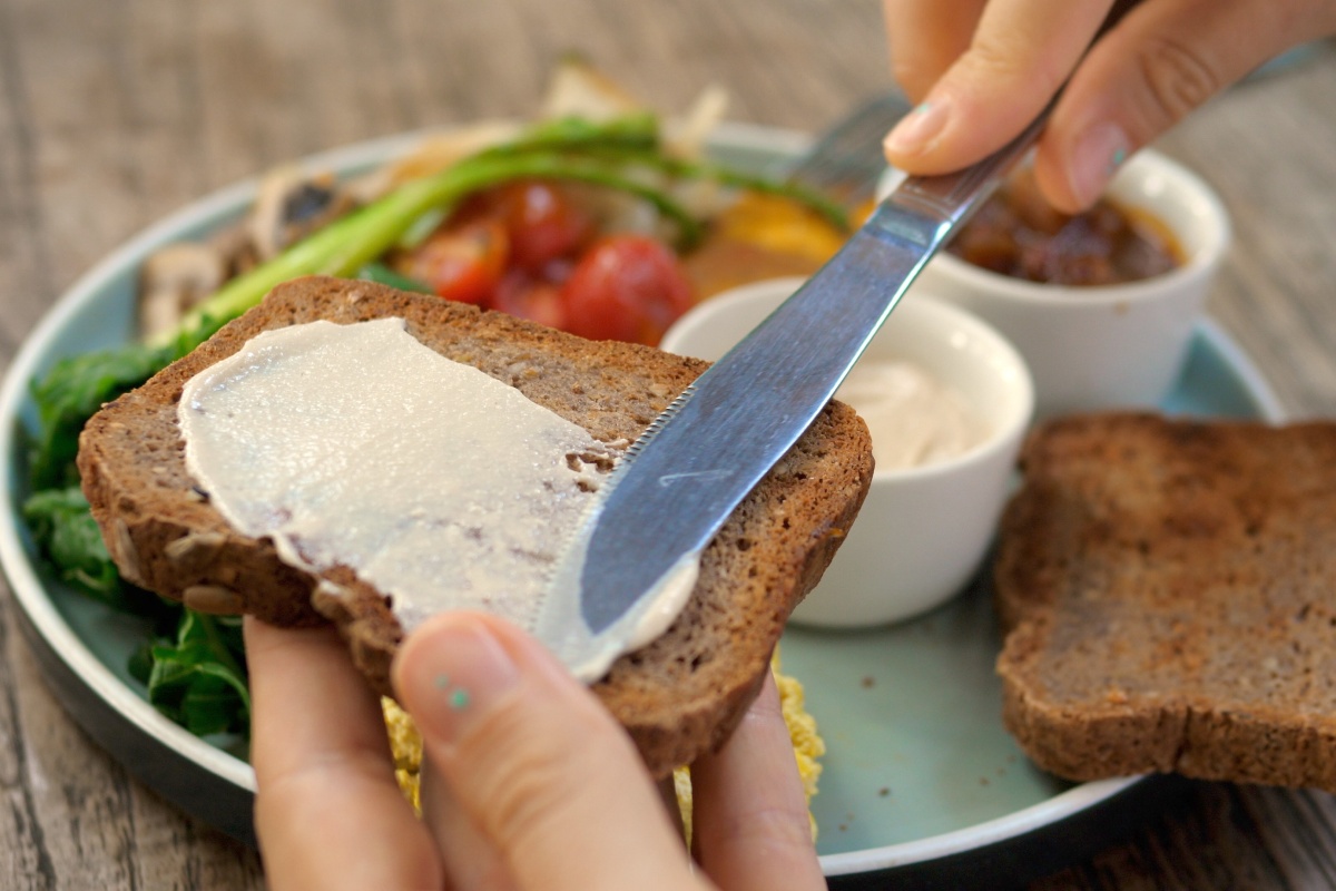 Person streicht Frischkäse auf eine Scheibe Vollkornbrot, mit frischem Gemüse und Dips im Hintergrund.