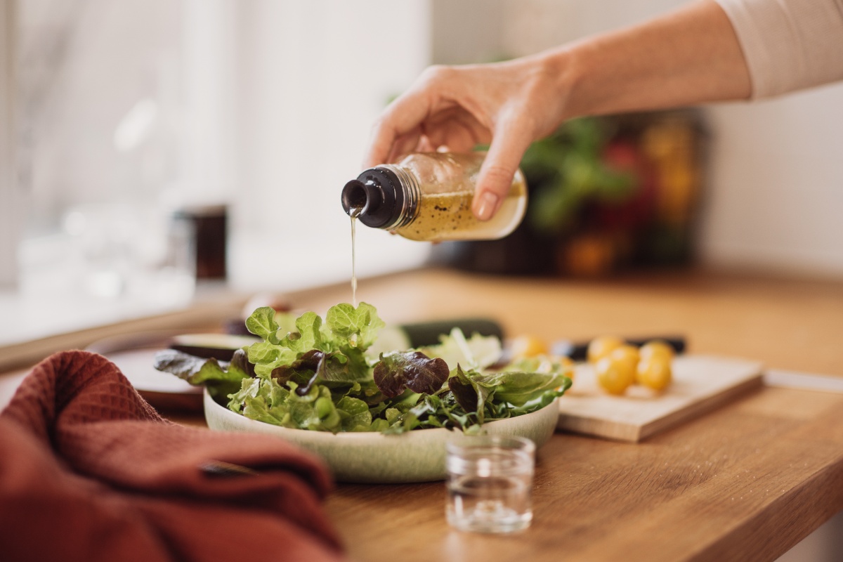 Eine Person gießt Vinaigrette aus einer Flasche über frischen grünen Salat in einer Schale auf einem Holztisch.