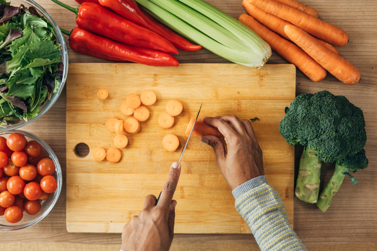 Person schneidet Karotten auf einem Holzbrett, umgeben von frischem Gemüse.