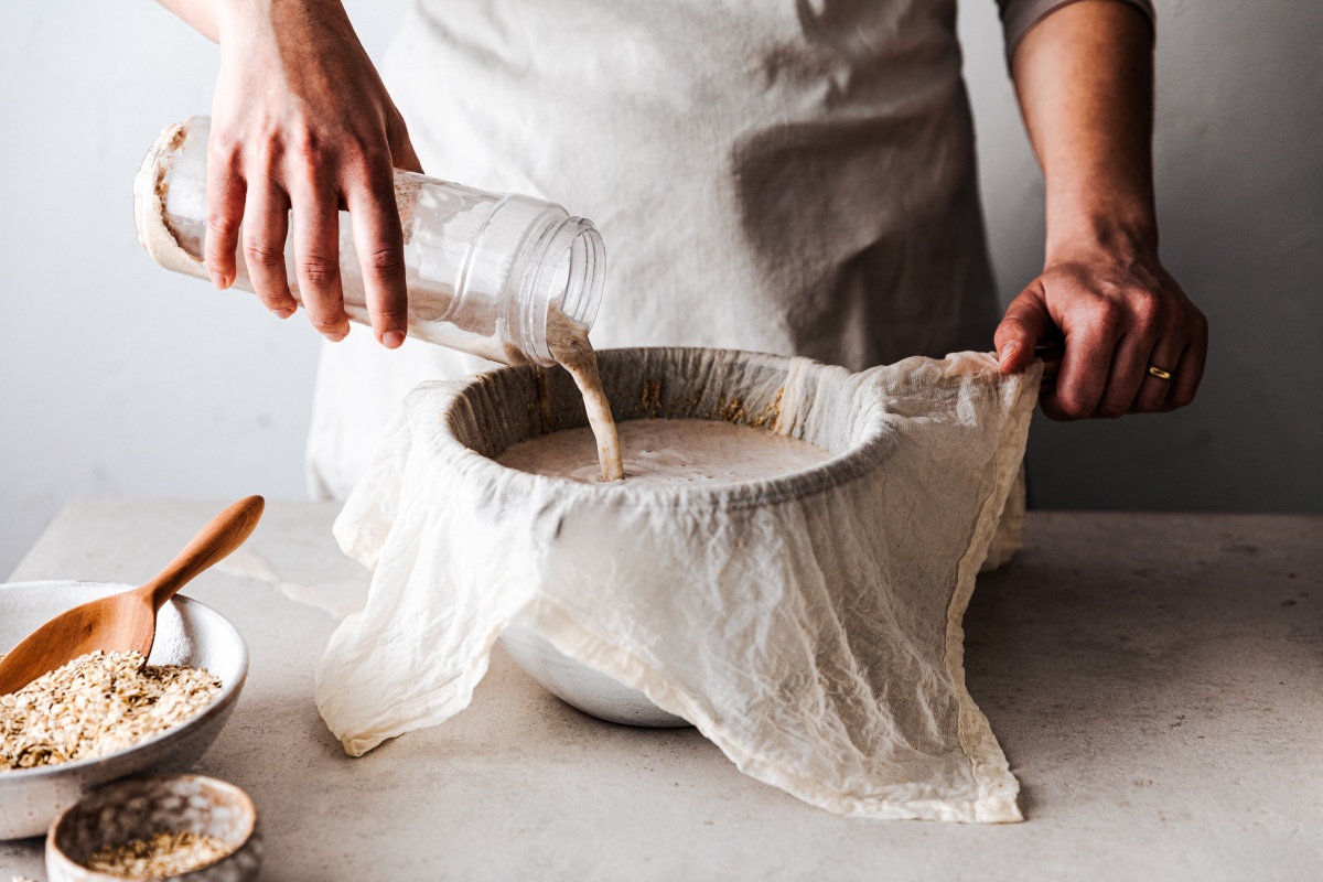 Person gießt selbstgemachte Hafermilch in eine mit Tuch ausgelegte Schüssel.