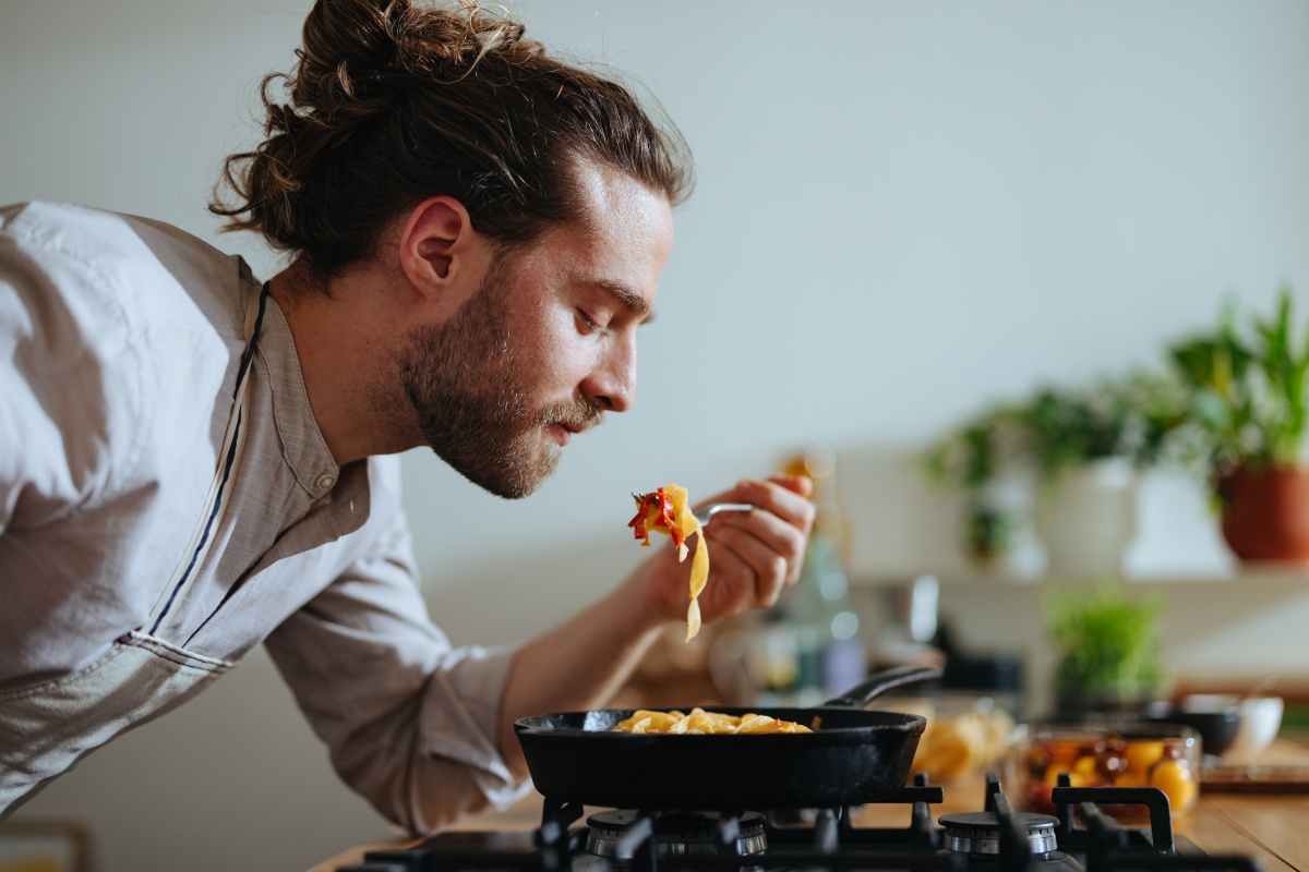 Mann mit Kochschürze probiert frisch gekochte Pasta direkt aus der Pfanne.