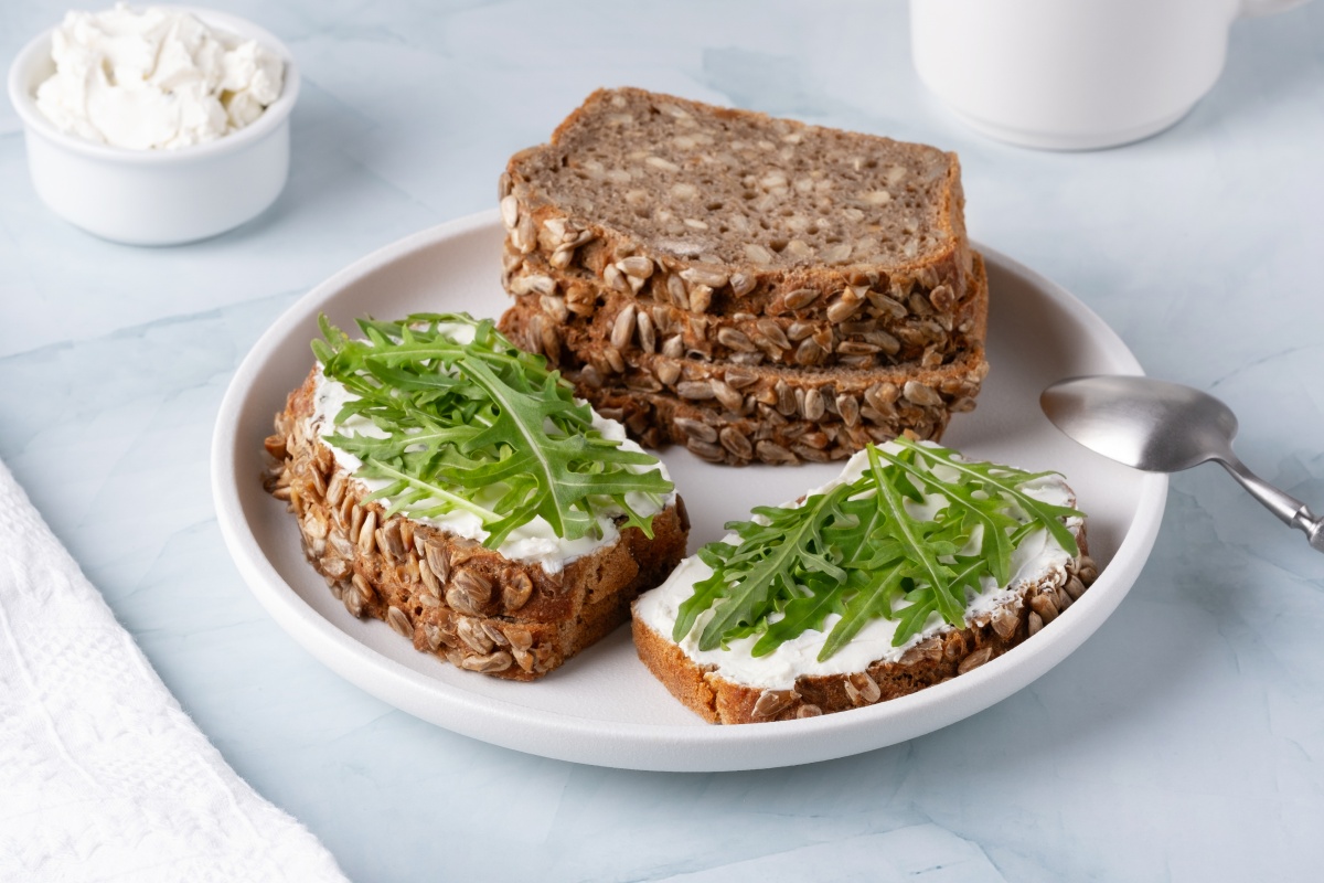 Vollkornbrot mit Sonnenblumenkernen, bestrichen mit Frischkäse und Rucola, auf einem Teller, daneben eine Schale Frischkäse.
