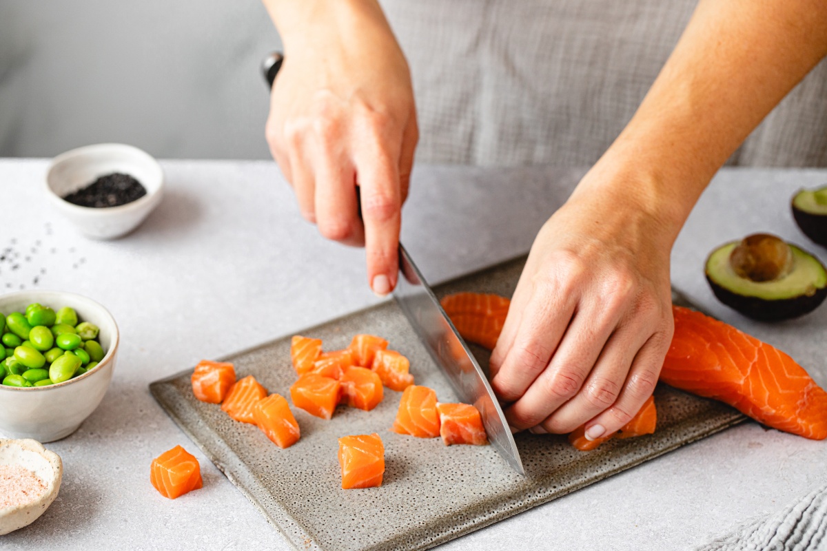 Person schneidet frischen Lachs in Würfel, daneben Schalen mit Edamame, Salz und eine halbe Avocado.