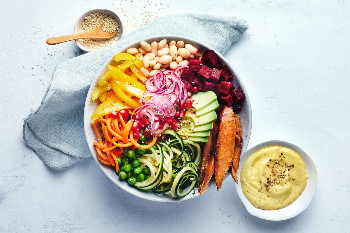 Bunte Buddha-Bowl mit Avocado, Süßkartoffel, Gurke, Paprika, Zwiebeln, Bohnen und Roter Bete, daneben eine Schale mit Dressing.