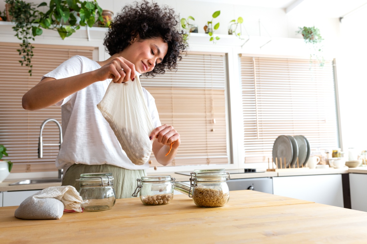 Frau füllt Hülsenfrüchte aus einem Stoffbeutel in Gläser in einer modernen hellen Küche.