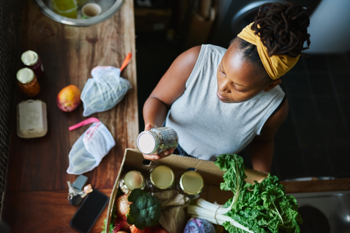 Eine Frau packt frische Lebensmittel wie Gemüse und Gläser mit Konserven aus einem Karton in einer Küche aus.