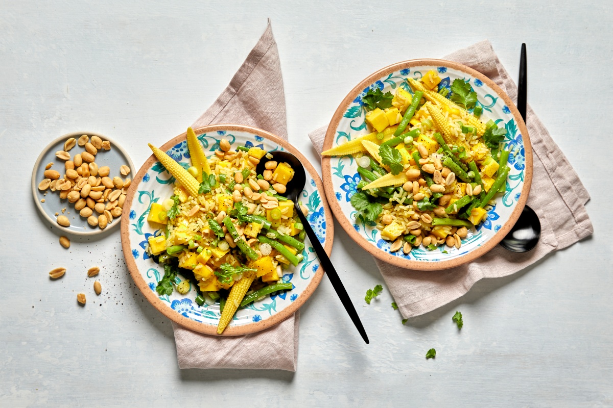 Reissalat mit Babymais, grünen Bohnen, Mango und Erdnüssen, serviert in zwei Schalen mit frischen Kräutern und Löffeln.