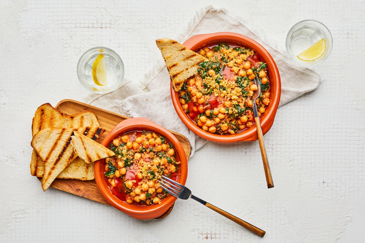 Spanische Kichererbsen mit Spinat und Tomaten, garniert mit geröstetem Brot, serviert in zwei Schalen auf einem Tisch mit Zitronenwasser.