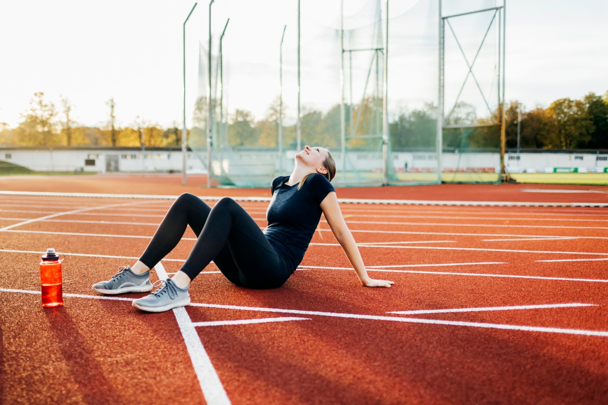 Frau in Sportkleidung sitzt auf einer Laufbahn und erholt sich nach einem intensiven Training, neben ihr eine Wasserflasche.