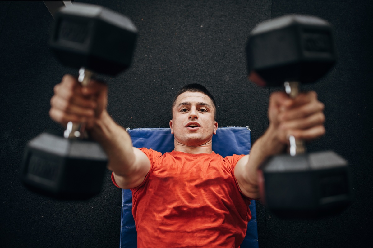 Mann in rotem Shirt liegt auf einer Matte und hebt Kurzhanteln während eines Krafttrainings im Fitnessstudio.