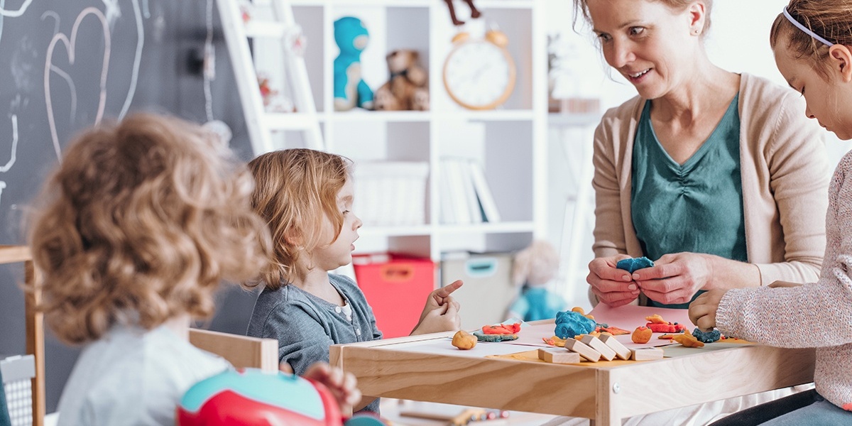 Eine Frau und drei Kleinkinder spielen mit Knete an einem Tisch.