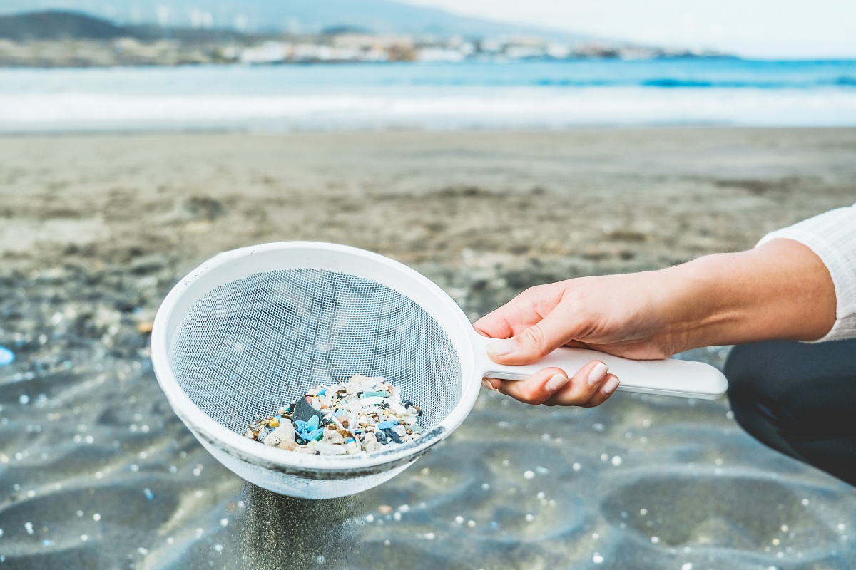 Hand mit Sieb voller Kunststoffteilchen am Strand.