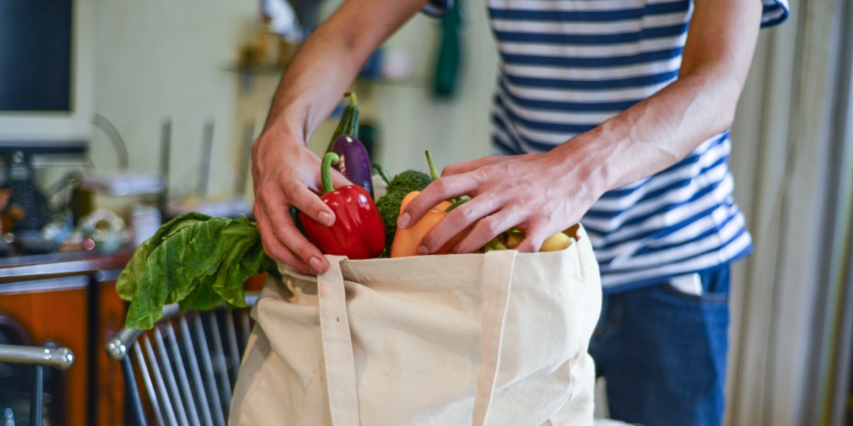 Ein Mann in gestreiftem Shirt packt Gemüse wie Paprika, Salat und Aubergine in eine beigefarbene Stofftasche.