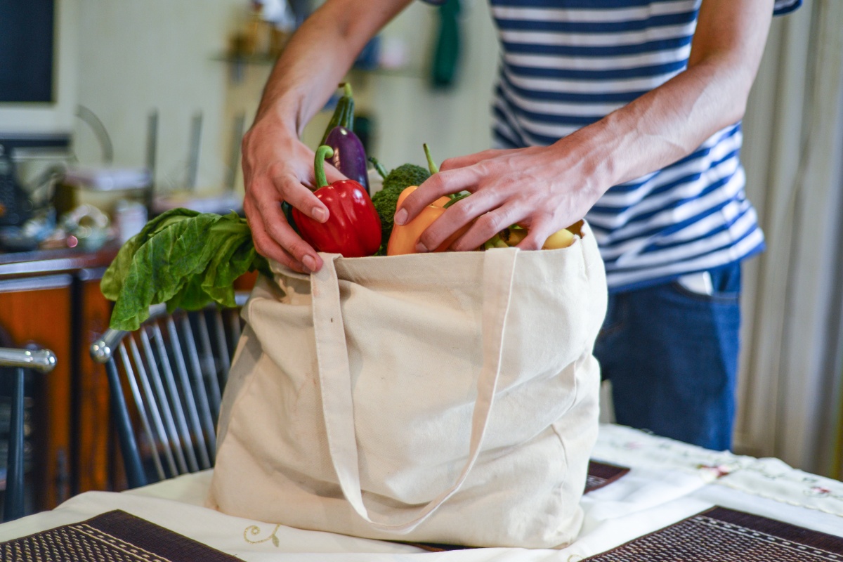 Ein Mann in gestreiftem Shirt packt Gemüse wie Paprika, Salat und Aubergine in eine beigefarbene Stofftasche.