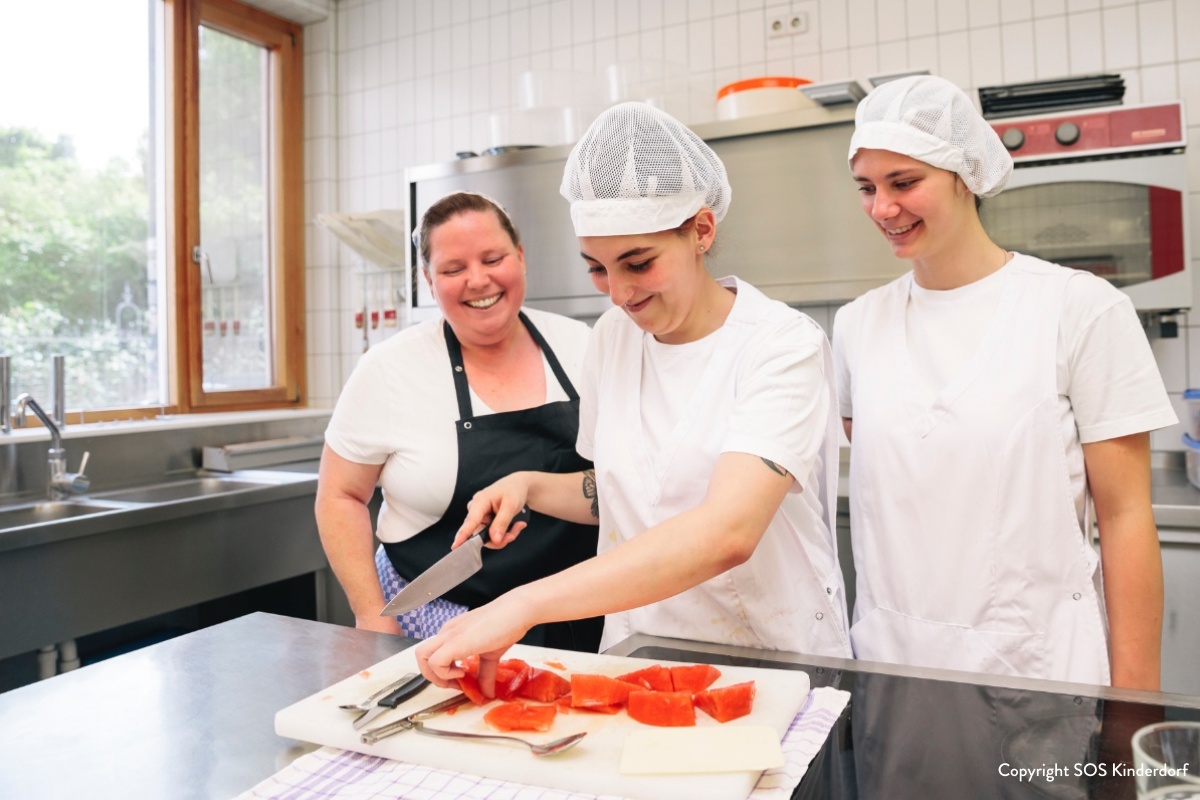 Eine lächelnde Köchin steht zusammen mit zwei Jugendlichen in einer Küche. Er davon schneidet eine Wassermelone.