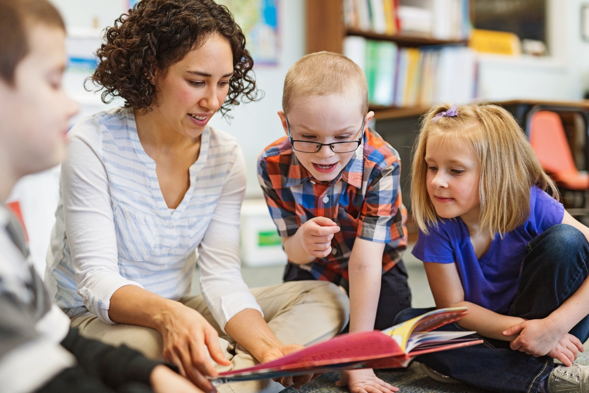 Eine Frau liest drei Kindern etwas aus einem Buch vor.