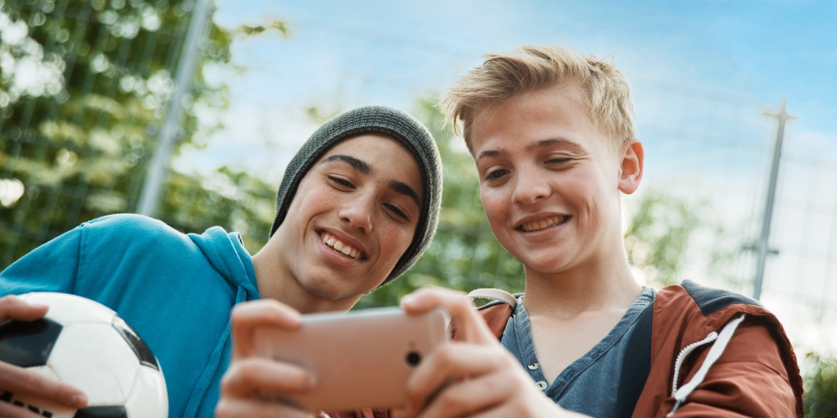 Zwei lächelnde Jungs, einer mit Fußball in der Hand, schauen auf ein Smartphone.