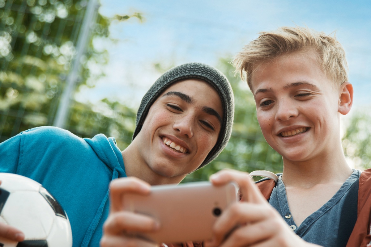 Zwei lächelnde Jungs, einer mit Fußball in der Hand, schauen auf ein Smartphone.