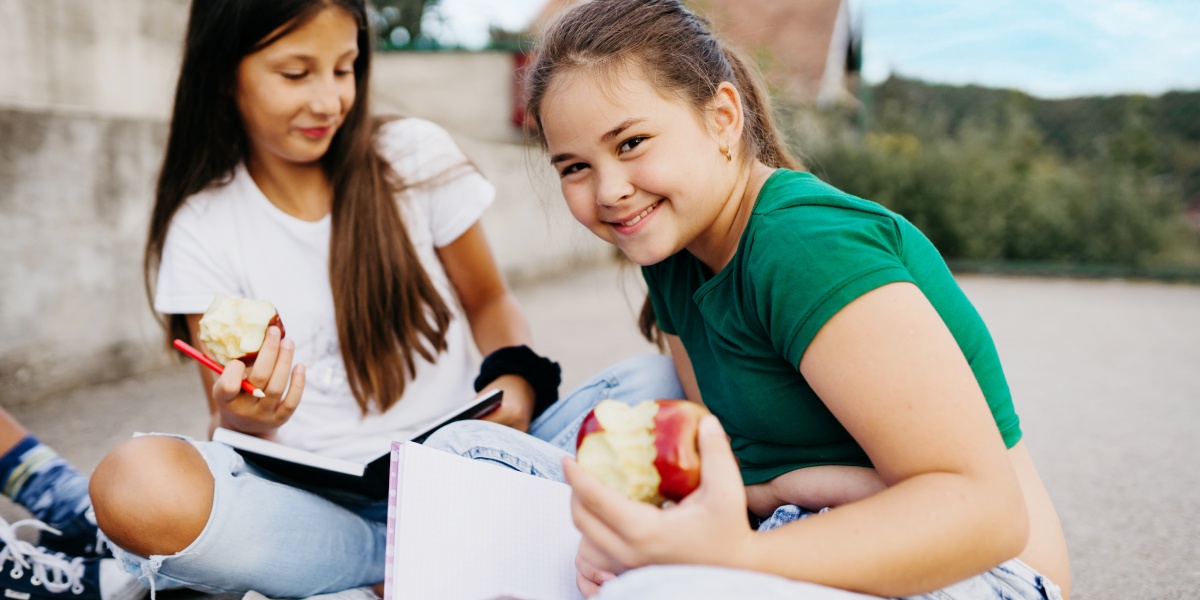 Zwei Mädchen sitzen auf dem Boden, essen einen Apfel und notieren etwas in Notizbüchern.
