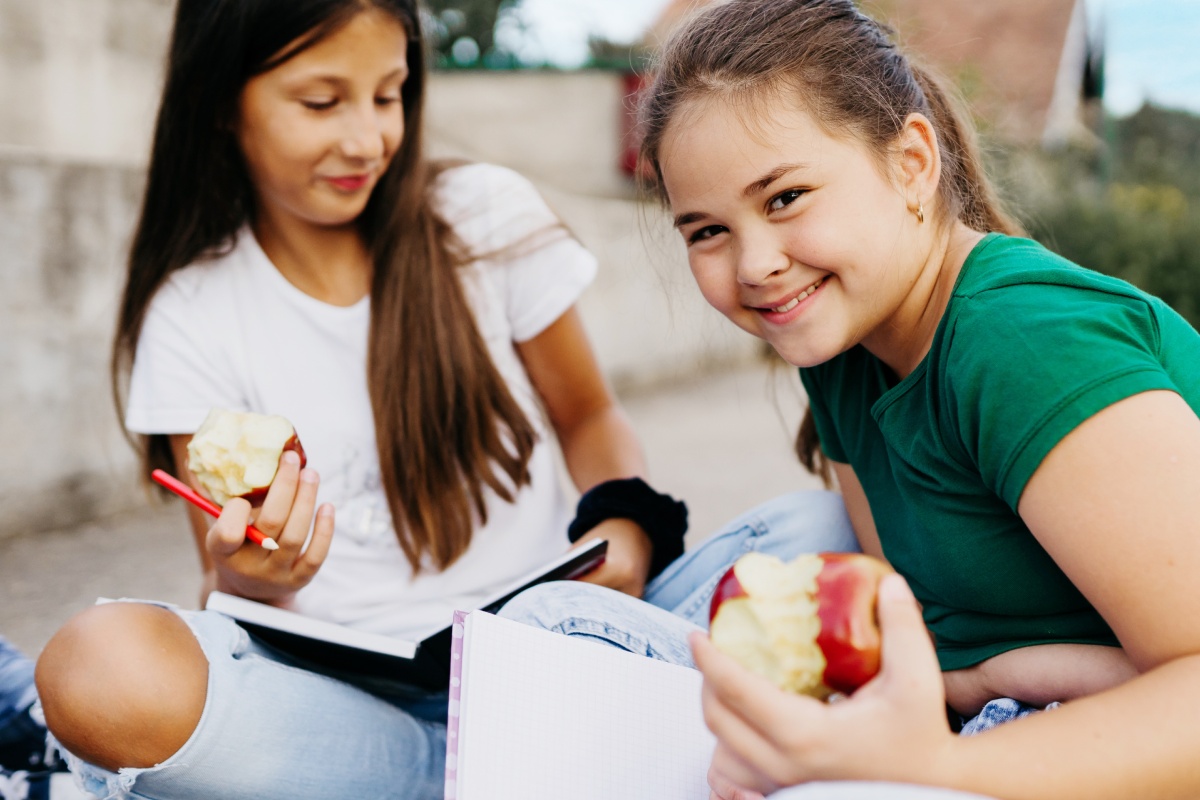 Zwei Mädchen sitzen auf dem Boden, essen einen Apfel und notieren etwas in Notizbüchern.