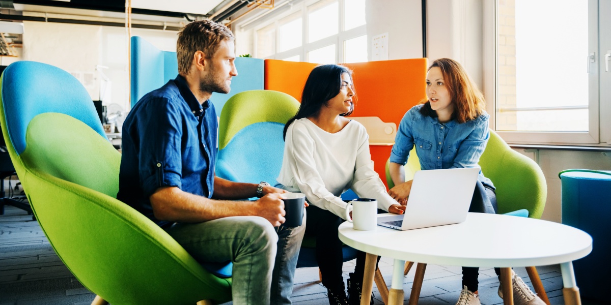 Drei Kollegen diskutieren in einem modernen Büro, während sie an einem Laptop arbeiten, helle Sessel im Hintergrund.