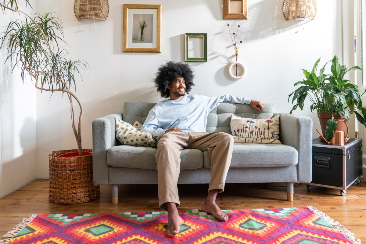 Eine Mann sitzt gemütlich auf dem grauen Sofa im Wohnzimmer. Vor ihm liegt ein bunter Teppich.