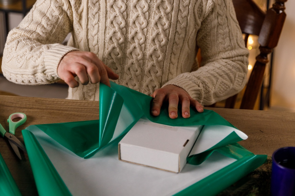 Person in Strickpullover verpackt eine kleine Box in grünes Geschenkpapier.