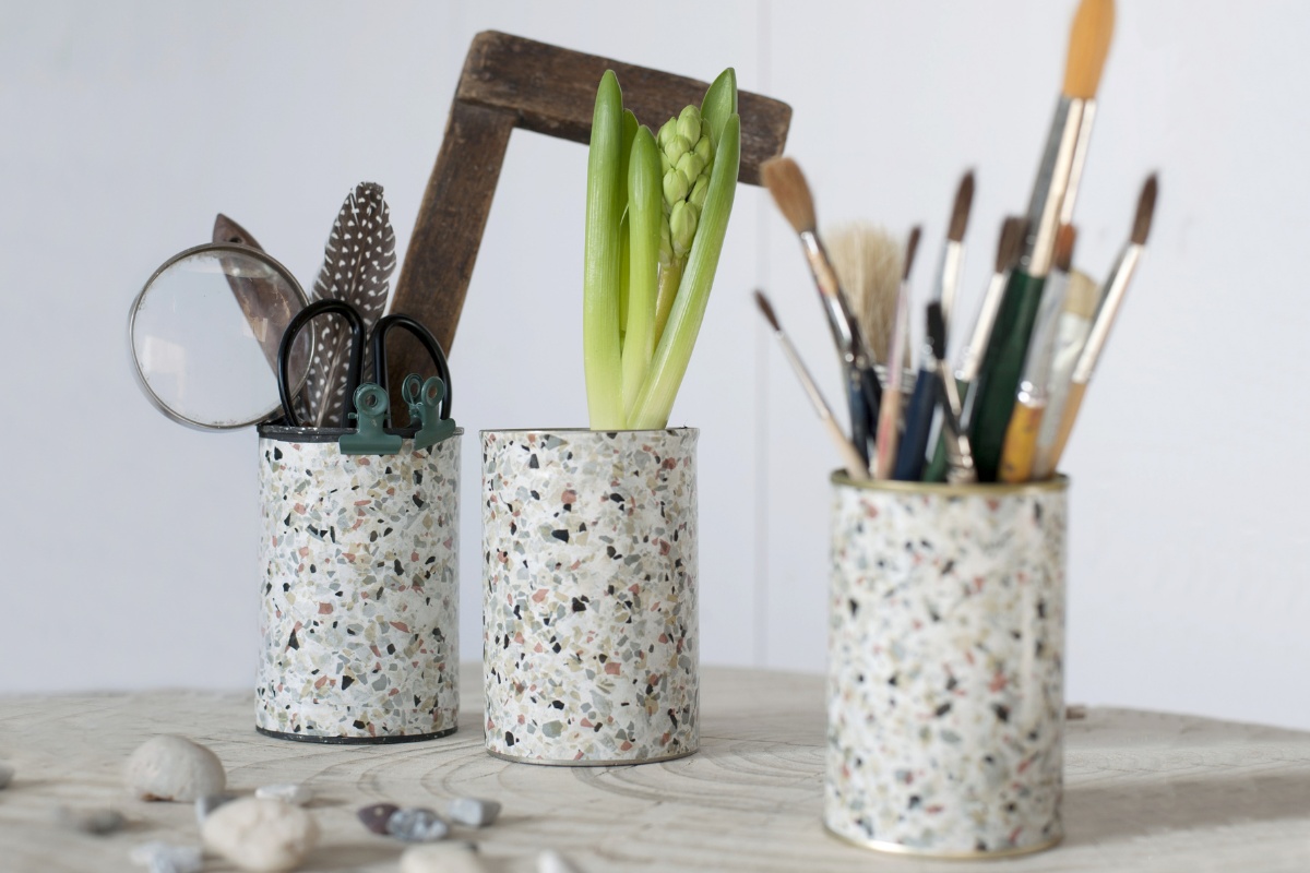 Auf einem Tisch stehen drei länglich runde Dosen im Terrazzo-Look, eine gefüllt mit Pinseln, eine bepflanzt und die dritte gefüllt mit Lupe, Feder, Schere und Schlosserwinkel.