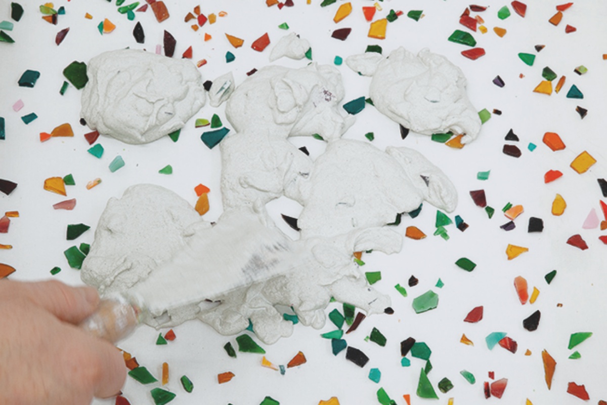 Person verteilt Sand-Zement-Mischung mit Spachtel auf einer Holzfaserplatte mit buntem Fliesenbruch.