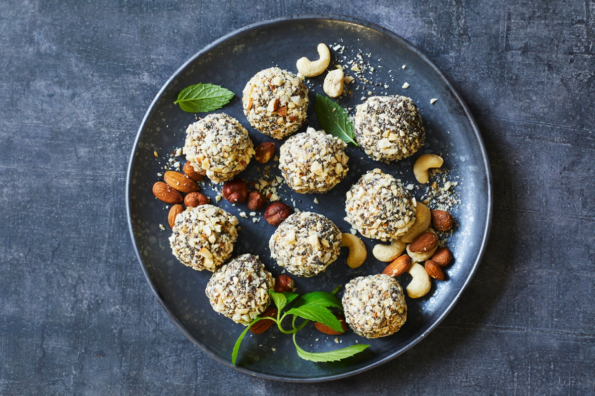 Energiekugeln mit Nusskruste auf dunklem Teller, garniert mit Mandeln, Cashews und Minzblättern, auf grauem Hintergrund.