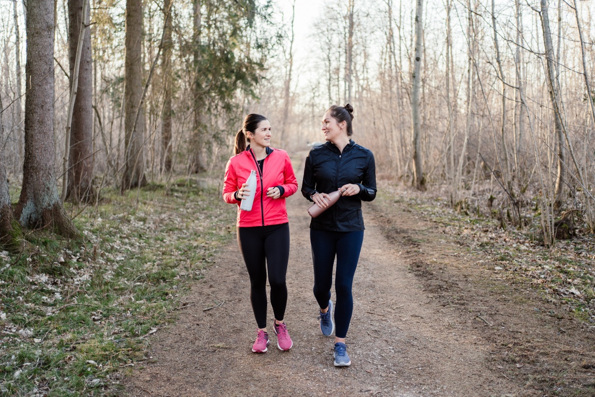 Zwei Frauen in Sportkleidung gehen mit Trinkflaschen auf einem Waldweg und unterhalten sich.