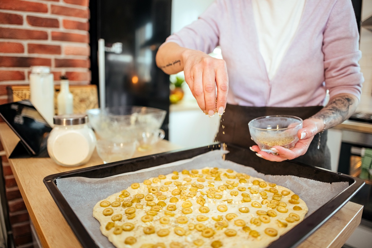 Person bestreut Teig mit Gewürzen und Olivenringen auf einem Backblech in der Küche.