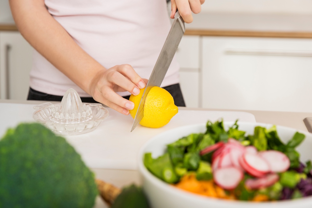 Person schneidet eine Zitrone auf einem Schneidebrett, daneben eine Glassaftpresse und eine Salatschüssel mit frischem Gemüse.