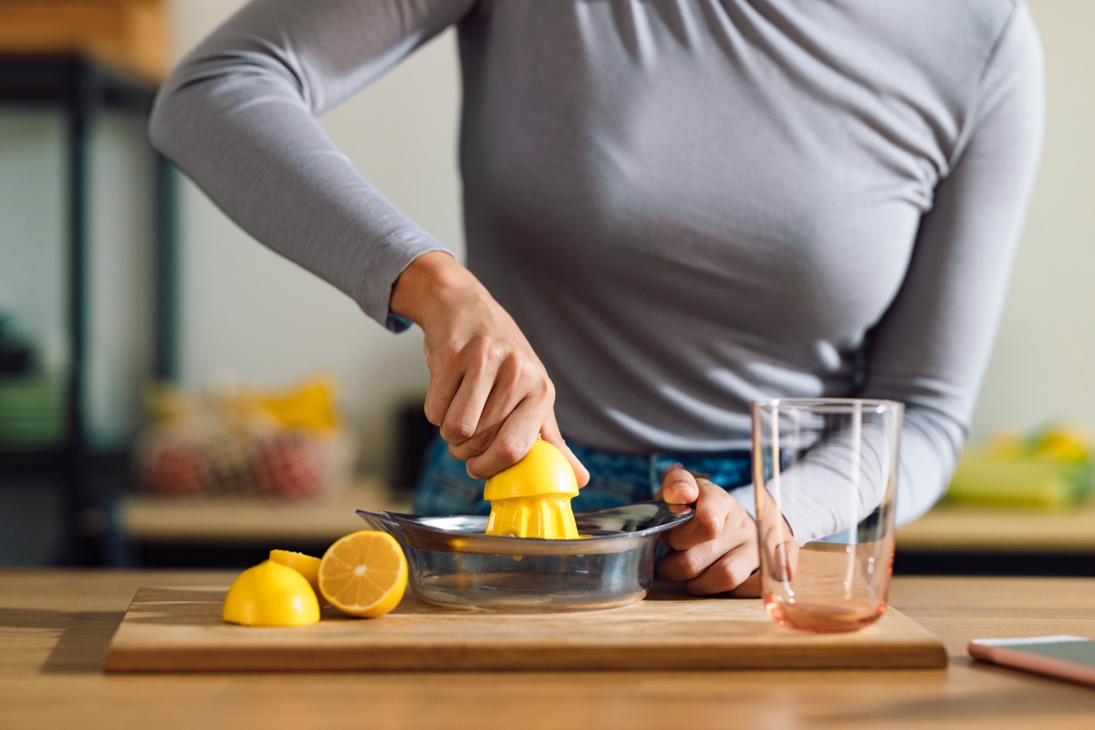Person presst eine Zitrone mit einer Handpresse, daneben halbe Zitronen und ein Glas auf einem Holztisch.
