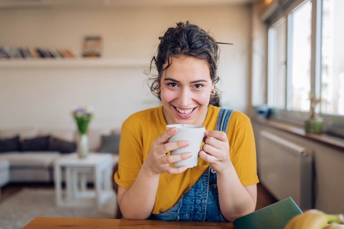 Junge Frau in gelbem Shirt und Latzhose hält eine Tasse und lächelt in die Kamera.