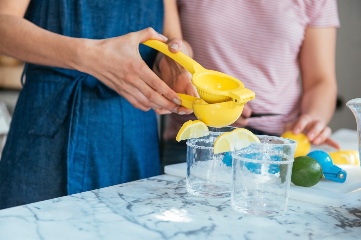 Frau mit blauer Küchenschürze presst Zitronensaft mit einer gelben Handpresse in Gläser.