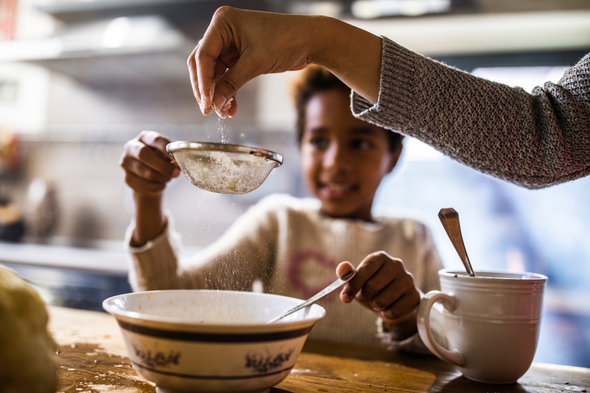 Erwachsene Person siebt Mehl in eine Schale, während ein Kind zuschaut und rührt.
