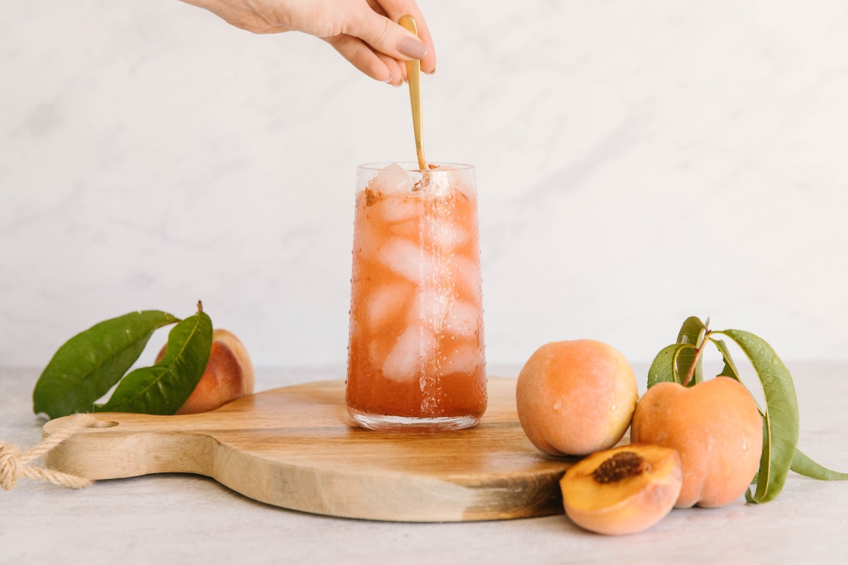 Eine Hand rührt einen Eistee um, daneben liegen Pfirsiche.