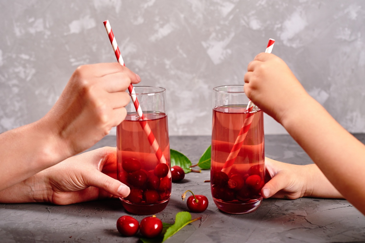 Zwei erwachsene Hände halten Kirsch-Eistee mit Strohalm, gegenüber zwei Kinderhände mit gleichem Getränk.