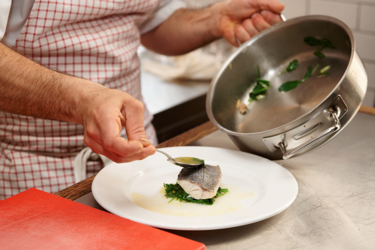 Ein Stück gedünsteter Fisch auf einem Teller wird mit Sud übergossen.